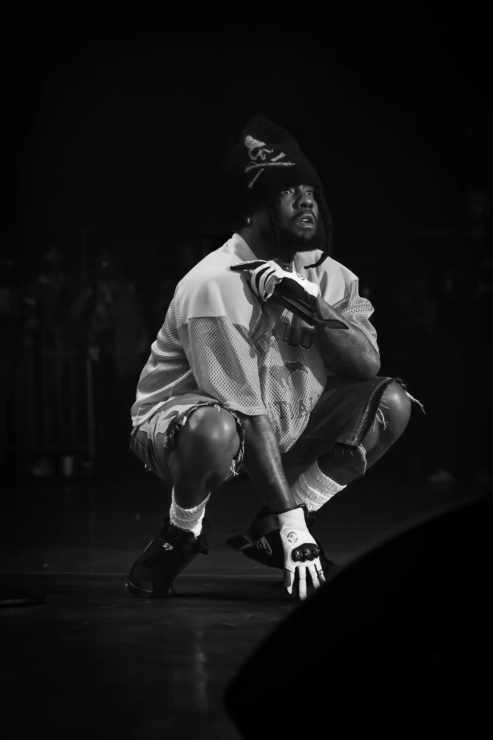 Wale crouching on stage holding a microphone at Roots Picnic 2025, black and white