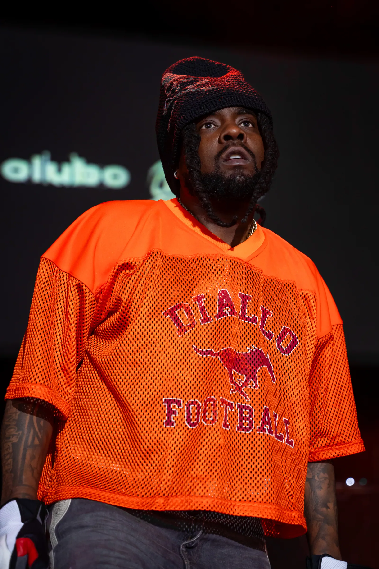 Wale close-up in orange Diallo Football jersey at Roots Picnic 2025, looking up at the crowd