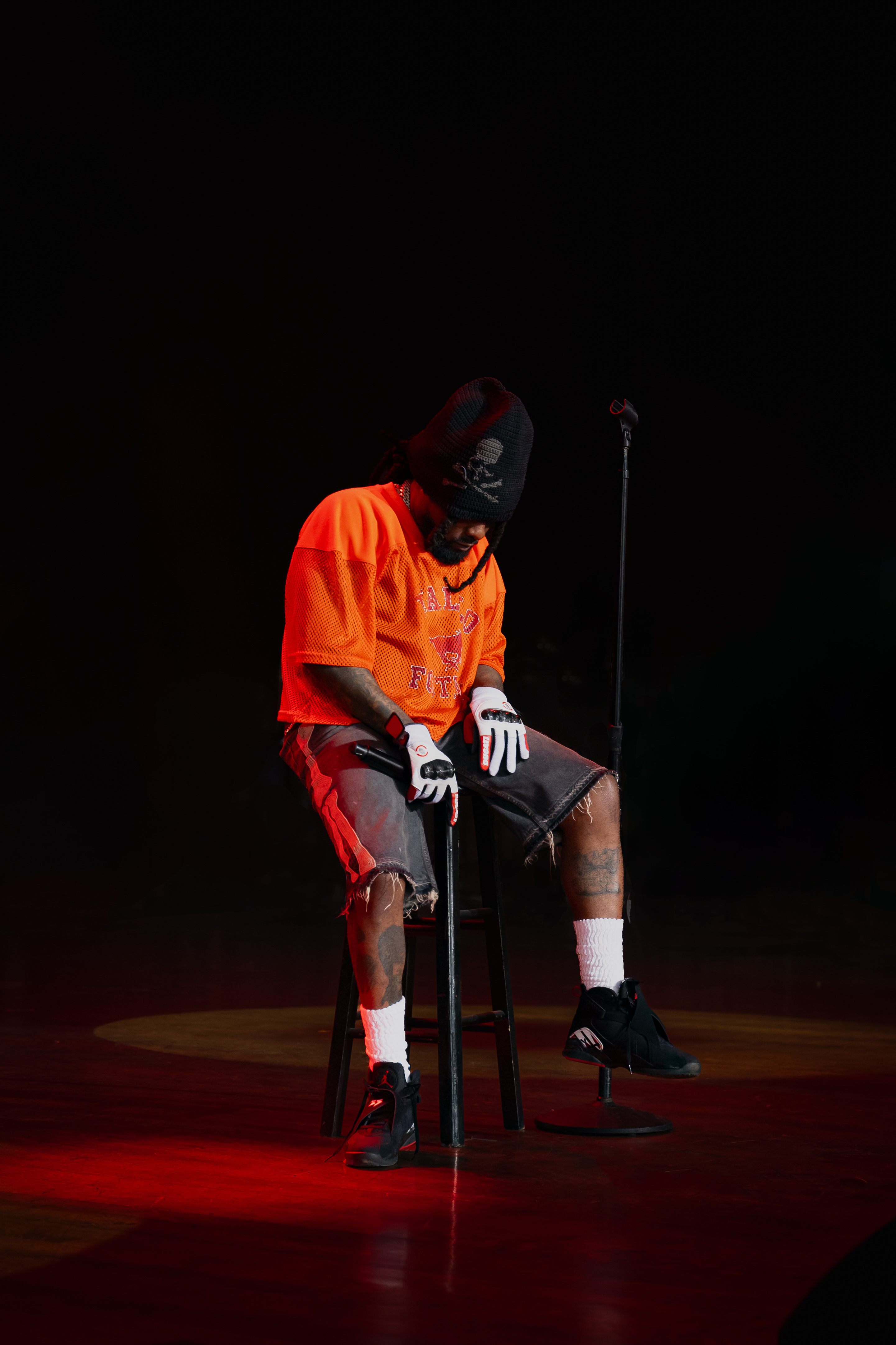 Wale seated on a stool on stage at Roots Picnic 2025, head down, dramatic red stage lighting
