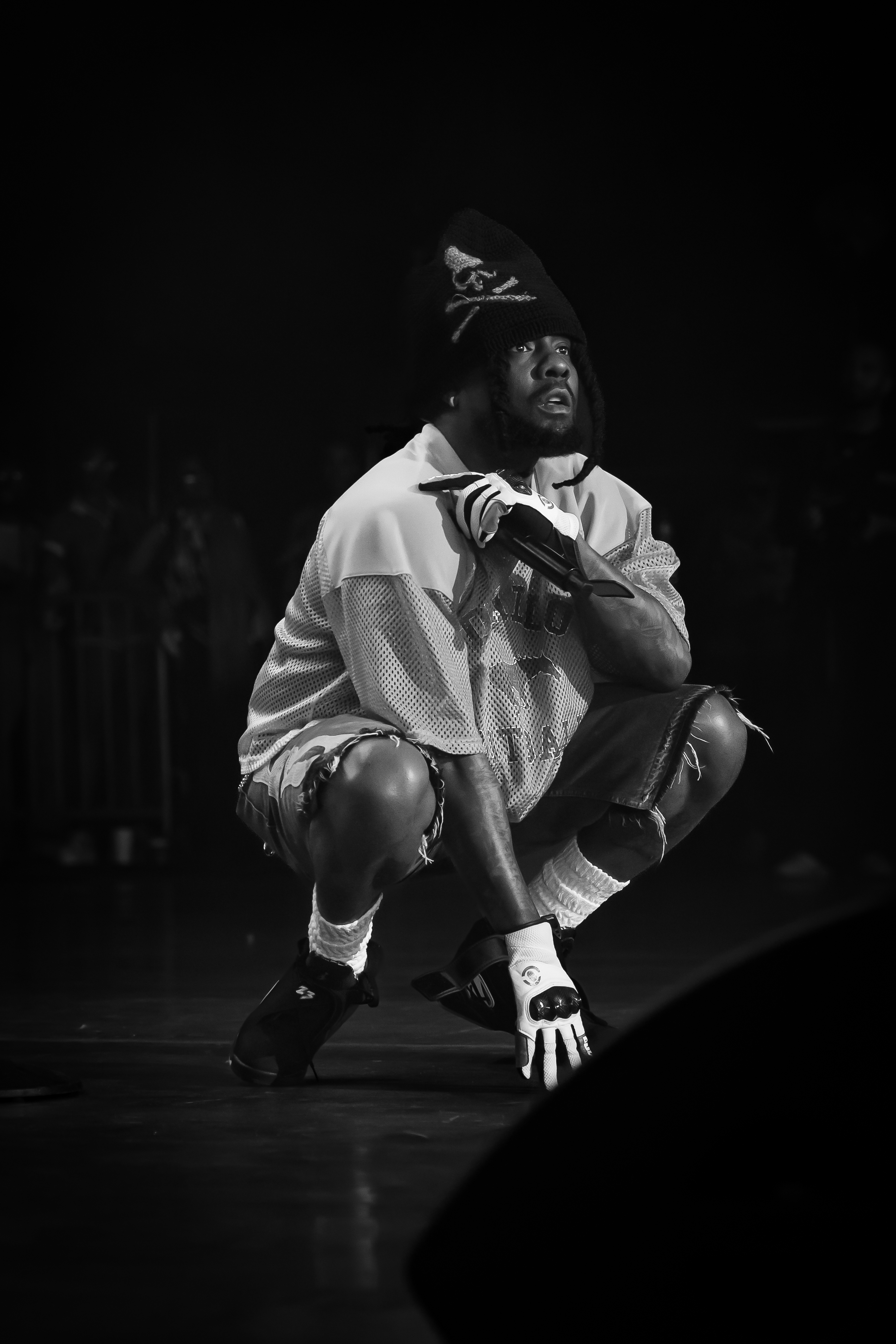 Wale crouching on stage holding a microphone at Roots Picnic 2025, black and white